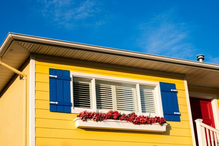 A yellow house with blue shudders and pink flowers below the window.
