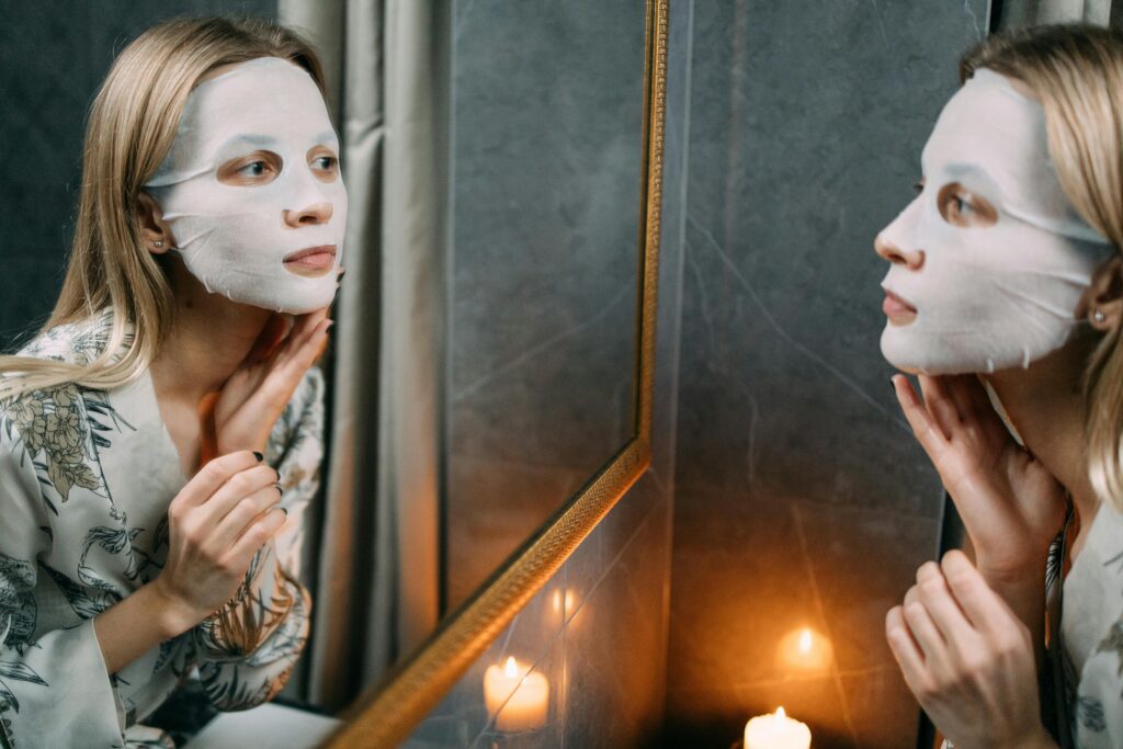 A woman with blond hair looks into a bathroom mirror with a face mask on to practice skincare.