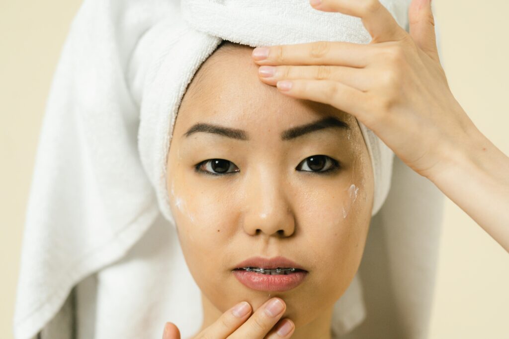 A woman has a towel around her head to dry her hair, and is rubbing lotion on her face.